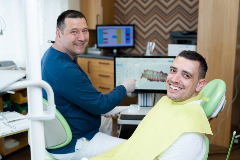 Egy mosolygó fogorvos a monitoron lévő fogászati felvételre mutat mellette egy mosolygó páciens.