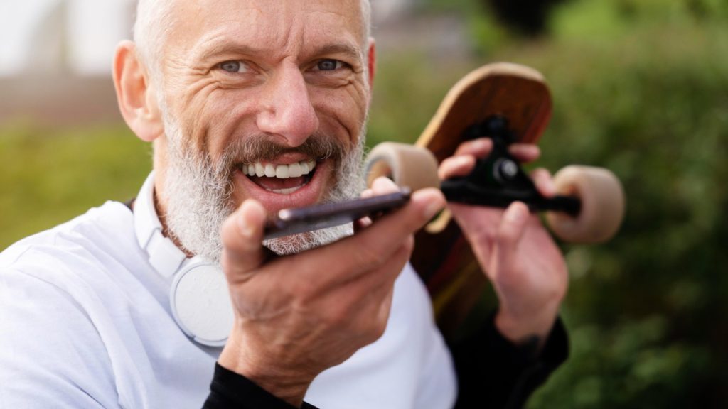 Sportos idős férfi mosolyog egyik kezében telefonnal a másikban gördeszkával.
